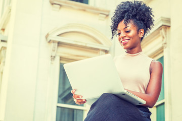 Way to Success. Young African American woman with afro hairstyle wearing sleeveless light color top, sitting by vintage office building in New York, looking down, working on laptop computer, smiling..
