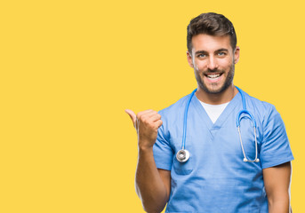 Young handsome doctor surgeon man over isolated background smiling with happy face looking and pointing to the side with thumb up.