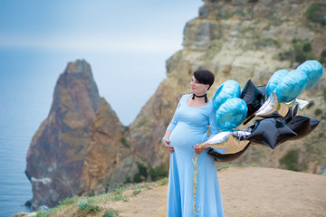 Pregnant woman posing on mountain coast line wearing blue long dress with air baloons.