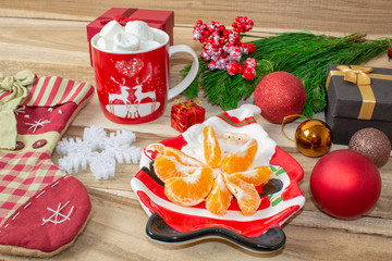 A set of Christmas decorations - a plate in the shape of Santa with tangerines, a sock for gifts, a mug of marshmallow, a branch of spruce and gifts with toys on a wooden table.