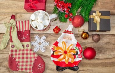 A set of Christmas decorations - a plate in the shape of Santa with tangerines, a sock for gifts, a mug of marshmallow, a branch of spruce and gifts with toys on a wooden table.