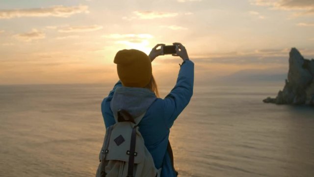young attractive girl hiking makes photo on smartphone. Shoots a beautiful sunset on a cape by the sea. Reached the goal. Crimea, Ukraine. The concept of freedom and unity with nature.