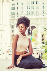 Fototapeta na wymiar Portrait of Young African American Female College Student, with afro hairstyle, wearing sleeveless top, skirt, bending legs, sitting on street by vintage style office building in New York, thinking..