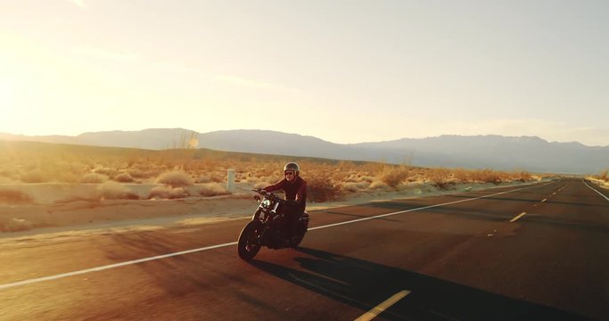 Happy man riding vintage black motorcycle down desert highway at sunset 