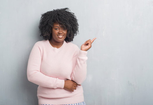 Young african american plus size woman over grey grunge wall wearing winter sweater with a big smile on face, pointing with hand and finger to the side looking at the camera.