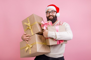 Happy man in Santa Claus hat holding a lot of Christmas gift boxes pinned to him on a pink background