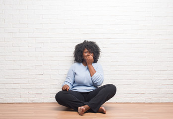 Young african american woman sitting on the floor at home looking stressed and nervous with hands on mouth biting nails. Anxiety problem.