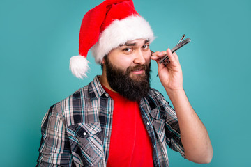 Bearded man in Santa Claus hat shaves a straight razor on a blue background