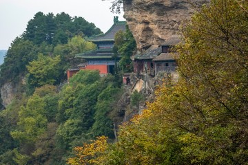 Red Taoist monastery and temple built on the side of a mountain with fall foliage and lush vegetation surrounding, in China