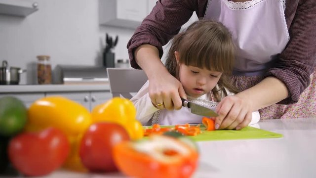 Engrossed little girl with down syndrome cutting red pepper on chopping boards with assistance of her loving mother while family preparing healthy food together in domestic kitchen.