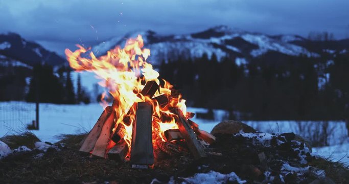 Burning Bonfire With Winter Mountains in Background. SLOW MOTION. Fairytale snow covered view with campfire burning outdoors at dusk. 