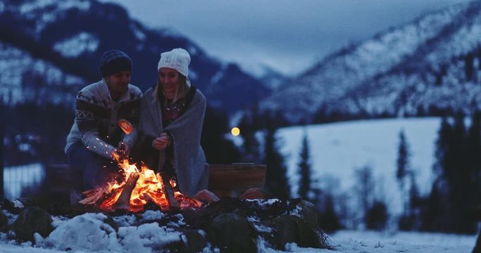Couple Sitting by the Burning Campfire With Winter Mountains in Background. SLOW MOTION. Fairytale snow covered view with fire burning outdoors at dusk. 
