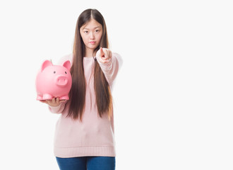 Young Chinese woman over isolated background holding piggy bank pointing with finger to the camera and to you, hand sign, positive and confident gesture from the front