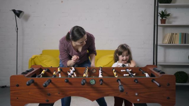Joyful happy mother and her excited laughing daughter with down syndrome playing table soccer in domestic room. Positive family with special needs child enjoying leisure playing football table soccer.