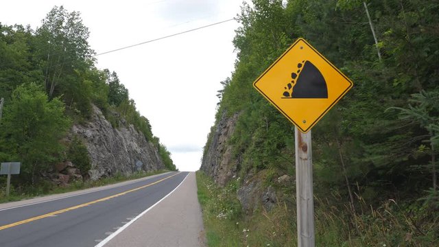 Yellow sign warning of falling rocks with rockcut and road ahead. Highway 35 near Miners Bay, Ontario, Canada.