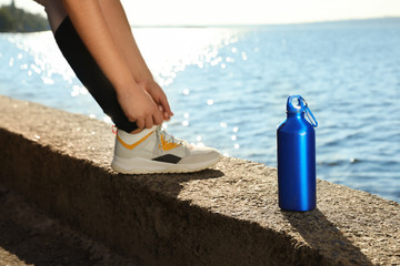 Young woman tying shoelaces near bottle of water at riverside on sunny day