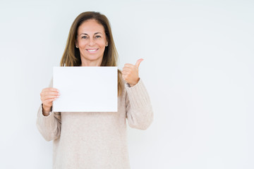 Middle age woman holding blank paper sheet over isolated background pointing and showing with thumb up to the side with happy face smiling