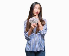 Young asian woman holding dollars over isolated background serious face thinking about question,...
