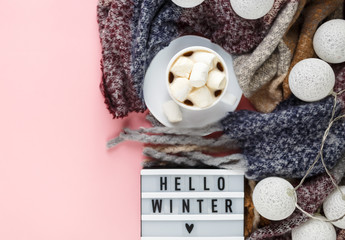 Warm, cozy winter clothing, scarf, lightbox and cup of coffee with white marshmallow as frame on pastel pink background. Christmas concept flat lay. hello winter title