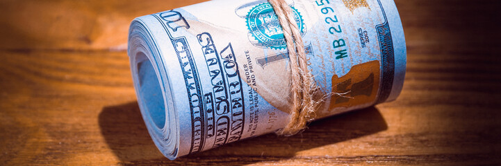 A roll of dollars on a wooden textured table in the dark illuminated by a ray of light.