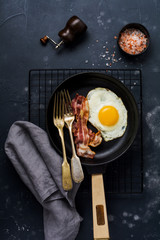 Traditional English breakfast with fried eggs and bacon in cast iron pan on dark concrete background. Top view.