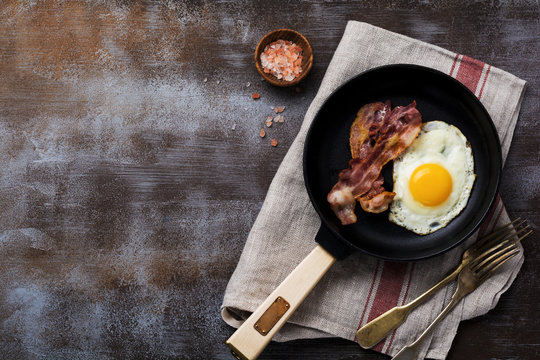 Traditional English breakfast with fried eggs and bacon in cast iron pan on dark concrete background. Top view.