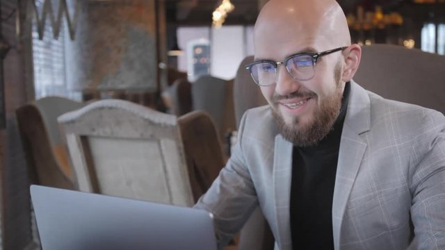 Carefully read and all of a sudden happily surprised. Portrait of young man with beard, wearing a suit in the glasses looking to the monitor laptop. Successful people, businessman in comfortable cafe