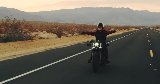 Happy man riding motorcycle at sunset celebrating with arms raised in air