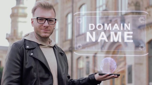 Smart young man with glasses shows a conceptual hologram Domain name. Student in casual clothes with future technology mobile screen on university background