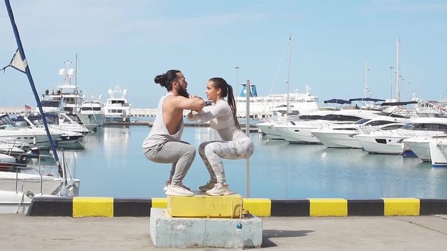 Fit young woman and athletic beared man box jumping in a crossfit style at the ocean pier with marinas. Female and male athlete is performing box jumps outdoor.