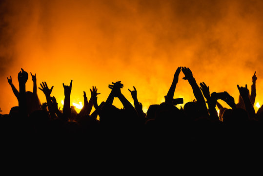 silhouettes of concert crowd in front of bright stage lights. Dancing people with hands on against stage light. Fans burn red flares at rock concert