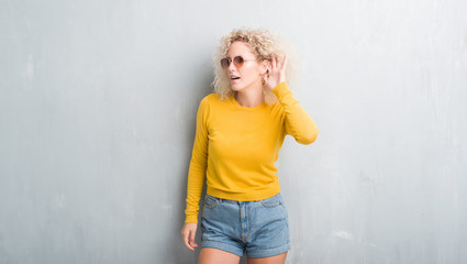 Young blonde woman with curly hair over grunge grey background smiling with hand over ear listening an hearing to rumor or gossip. Deafness concept.