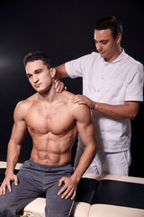 Fototapeta na wymiar two young man, 20-29 years old, sports physiotherapy indoors in studio, photo shoot. Physiotherapist massaging muscular patient shoulder with his hands, while he is sitting on bed.