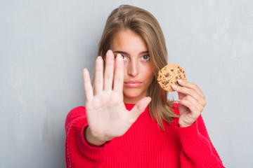 Beautiful young woman over grunge grey wall eating chocolate chip cooky with open hand doing stop sign with serious and confident expression, defense gesture