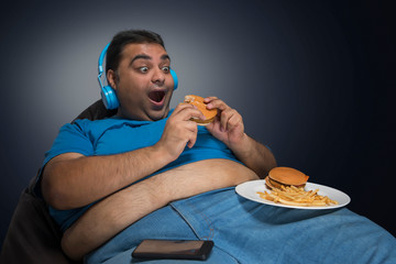 Excited obese man with belly visible out of his shirt eating burger while listening to music with a...