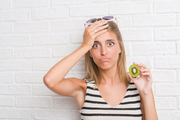 Beautiful young woman over white brick wall eating green kiwi stressed with hand on head, shocked with shame and surprise face, angry and frustrated. Fear and upset for mistake.