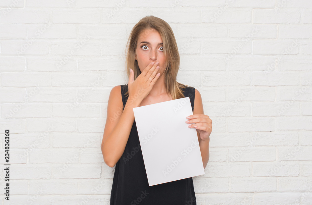 Wall mural Beautiful young woman over white brick wall holding blank paper sheet cover mouth with hand shocked with shame for mistake, expression of fear, scared in silence, secret concept