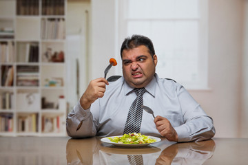 Overweight man sitting at dining table with plate full of vegetable salad holding a tomato slice...