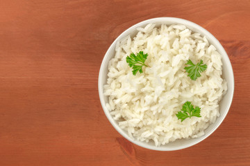 A bowl of cooked white long grain Basmati rice, garnished with parsley leaves, shot from above on a rustic wooden background with copy space