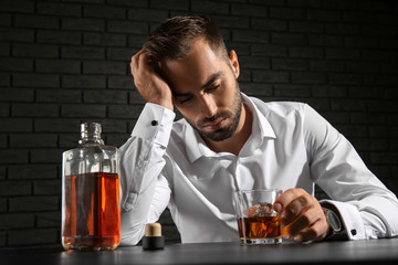 Drunk man with glass and bottle of whiskey sitting at table on dark background