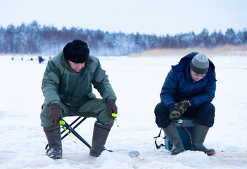 Fototapeta na wymiar Two caucasian man, old and young fisherman, are sitting on ice near their winter holes and fishing a smelt with several rods.