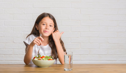 Young hispanic kid sitting on the table eating healthy salad pointing and showing with thumb up to the side with happy face smiling