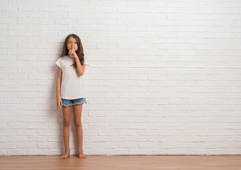Young hispanic kid stading over white brick wall asking to be quiet with finger on lips. Silence and secret concept.
