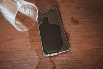 Smartphone wet by accident on wooden table. Glass of water spilled.