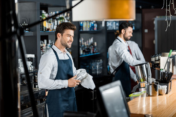 handsome smiling barman rubbing up glass with cloth at workplace