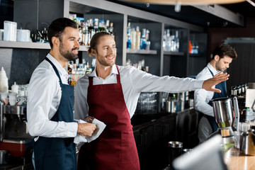 cheerful smiling barmen gesturing at workplace while coworker looking aside