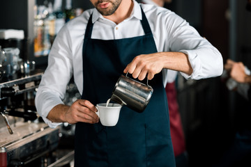 cropped view of barista in apron pouring milk to coffee at workplace
