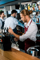 barman in apron pouring beer in glass behind wooden counter at bar