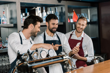 handsome adult barmen smiling and using smartphone at workplace
