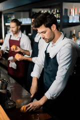handsome barman in apron slicing grapefruit with knife at counter